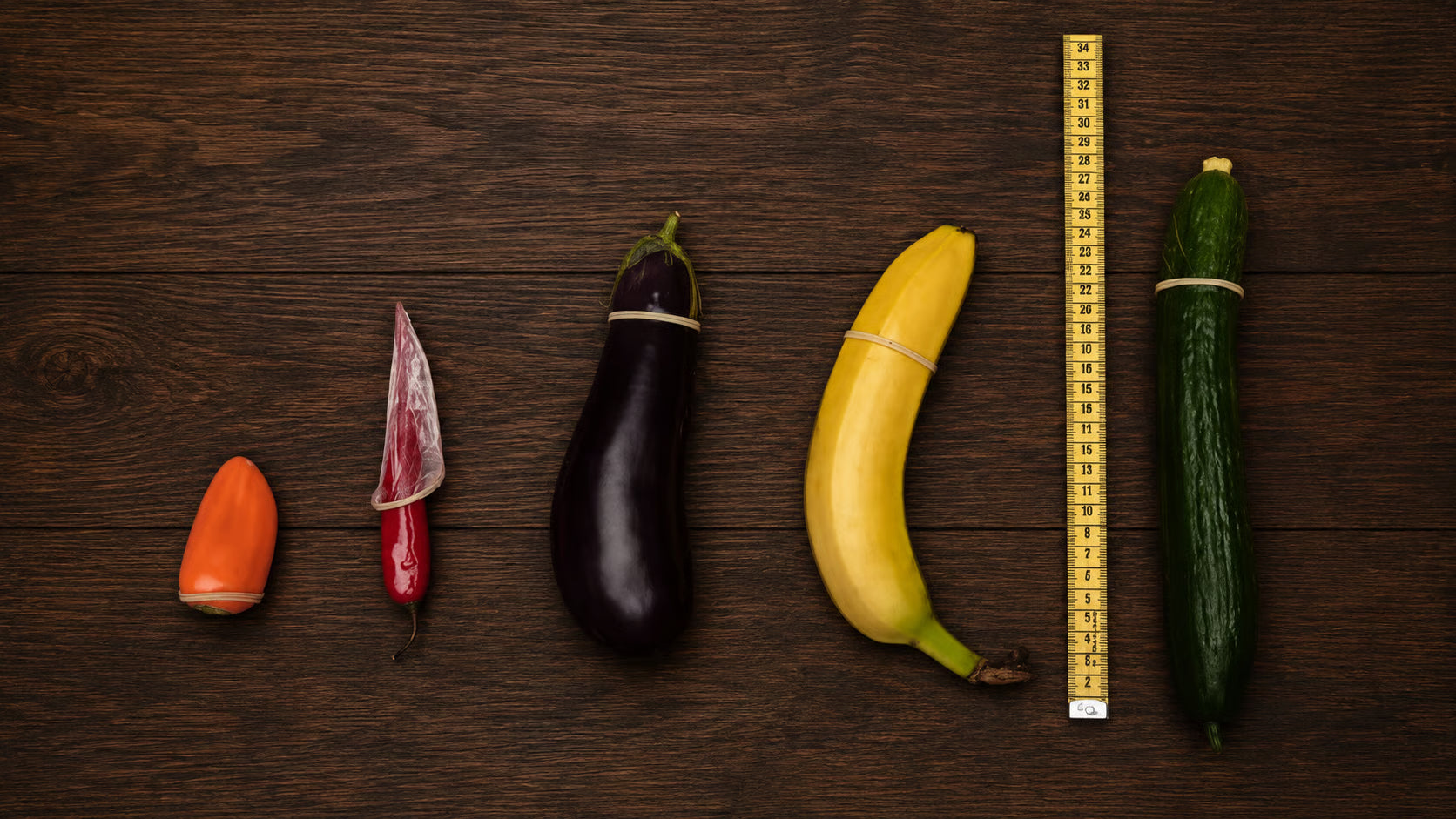 An overhead view of various vegetables and a banana arranged by size, with a condom on a chili.