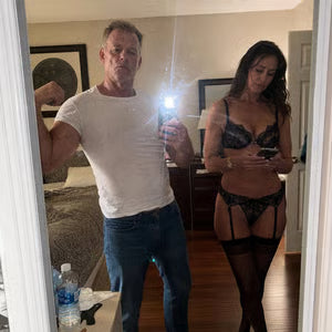 A man flexes for a mirror selfie as a woman in lingerie stands next to him looking at her phone.